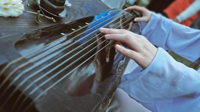A close-up shot captures a person playing a stringed musical instrument. The individual's hands gently press on the strings, which are stretched across a polished wooden surface. Soft fabric, possibly a traditional garment, covers the person's arms. A small flower and an incense holder are visible on the side of the instrument.