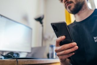 A person is sitting at a desk, holding a smartphone in their hand. In front of them is a computer monitor and a keyboard. The scene appears to be in a home or office setting, with the focus on the individual's hand and the smartphone.