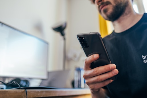 A person is sitting at a desk, holding a smartphone in their hand. In front of them is a computer monitor and a keyboard. The scene appears to be in a home or office setting, with the focus on the individual's hand and the smartphone.