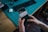 A pair of hands is holding a smartphone displaying notifications on its screen. The background consists of a teal cutting mat on a wooden desk, with a black keyboard and a white charging case visible. The setting suggests a workspace or desk environment.