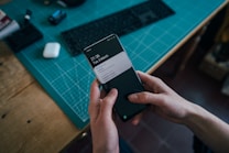 A pair of hands is holding a smartphone displaying notifications on its screen. The background consists of a teal cutting mat on a wooden desk, with a black keyboard and a white charging case visible. The setting suggests a workspace or desk environment.