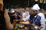A group of people is gathered around a food stall at what appears to be a festive outdoor event. The foreground shows a person in a traditional vest and white hat serving food. The background is decorated with colorful hanging ornaments and lights, creating a vibrant atmosphere.