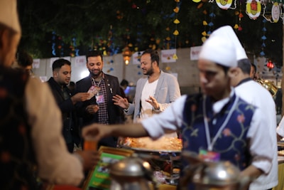 A group of people is gathered around a food stall at what appears to be a festive outdoor event. The foreground shows a person in a traditional vest and white hat serving food. The background is decorated with colorful hanging ornaments and lights, creating a vibrant atmosphere.