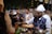 A group of people is gathered around a food stall at what appears to be a festive outdoor event. The foreground shows a person in a traditional vest and white hat serving food. The background is decorated with colorful hanging ornaments and lights, creating a vibrant atmosphere.