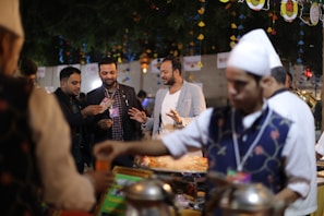 A group of people is gathered around a food stall at what appears to be a festive outdoor event. The foreground shows a person in a traditional vest and white hat serving food. The background is decorated with colorful hanging ornaments and lights, creating a vibrant atmosphere.