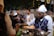 A group of people is gathered around a food stall at what appears to be a festive outdoor event. The foreground shows a person in a traditional vest and white hat serving food. The background is decorated with colorful hanging ornaments and lights, creating a vibrant atmosphere.