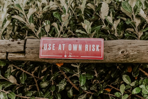 A red warning sign with white letters, reading 'USE AT OWN RISK', is attached to an aged wooden log. The background consists of dense green foliage.