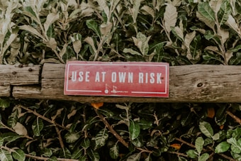 A red warning sign with white letters, reading 'USE AT OWN RISK', is attached to an aged wooden log. The background consists of dense green foliage.