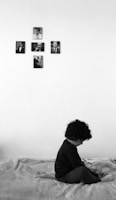 A young child sits on a bed with a pensive posture, looking down. The room is minimally decorated with five black and white photos arranged in a cross pattern on the wall above the child. The setting conveys a quiet, intimate atmosphere.