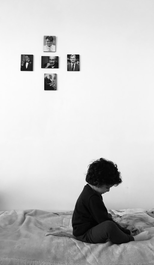 A young child sits on a bed with a pensive posture, looking down. The room is minimally decorated with five black and white photos arranged in a cross pattern on the wall above the child. The setting conveys a quiet, intimate atmosphere.