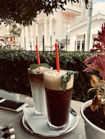 Two tall iced coffee drinks with red straws and mint leaves on top sit on a tray with a napkin. They are placed on an outdoor table next to a smartphone. In the background, a white building with a veranda and some greenery can be seen, along with blurred pedestrians.