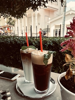 Two tall iced coffee drinks with red straws and mint leaves on top sit on a tray with a napkin. They are placed on an outdoor table next to a smartphone. In the background, a white building with a veranda and some greenery can be seen, along with blurred pedestrians.