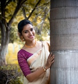A woman wearing a traditional sari is standing outdoors next to a tree with a background of blurred green and yellow foliage. She is smiling and looking at the camera, accessorized with gold jewelry including a necklace and bangles.
