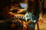 A young girl in a blue dress is sitting opposite an adult at a small chessboard, seemingly engaged in a game of chess. The focus is on the girl’s hands, which are expressive, as she gestures toward the chess pieces. The background is blurry, with a laptop visible in the distance.