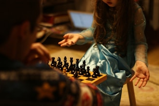 A young girl in a blue dress is sitting opposite an adult at a small chessboard, seemingly engaged in a game of chess. The focus is on the girl’s hands, which are expressive, as she gestures toward the chess pieces. The background is blurry, with a laptop visible in the distance.