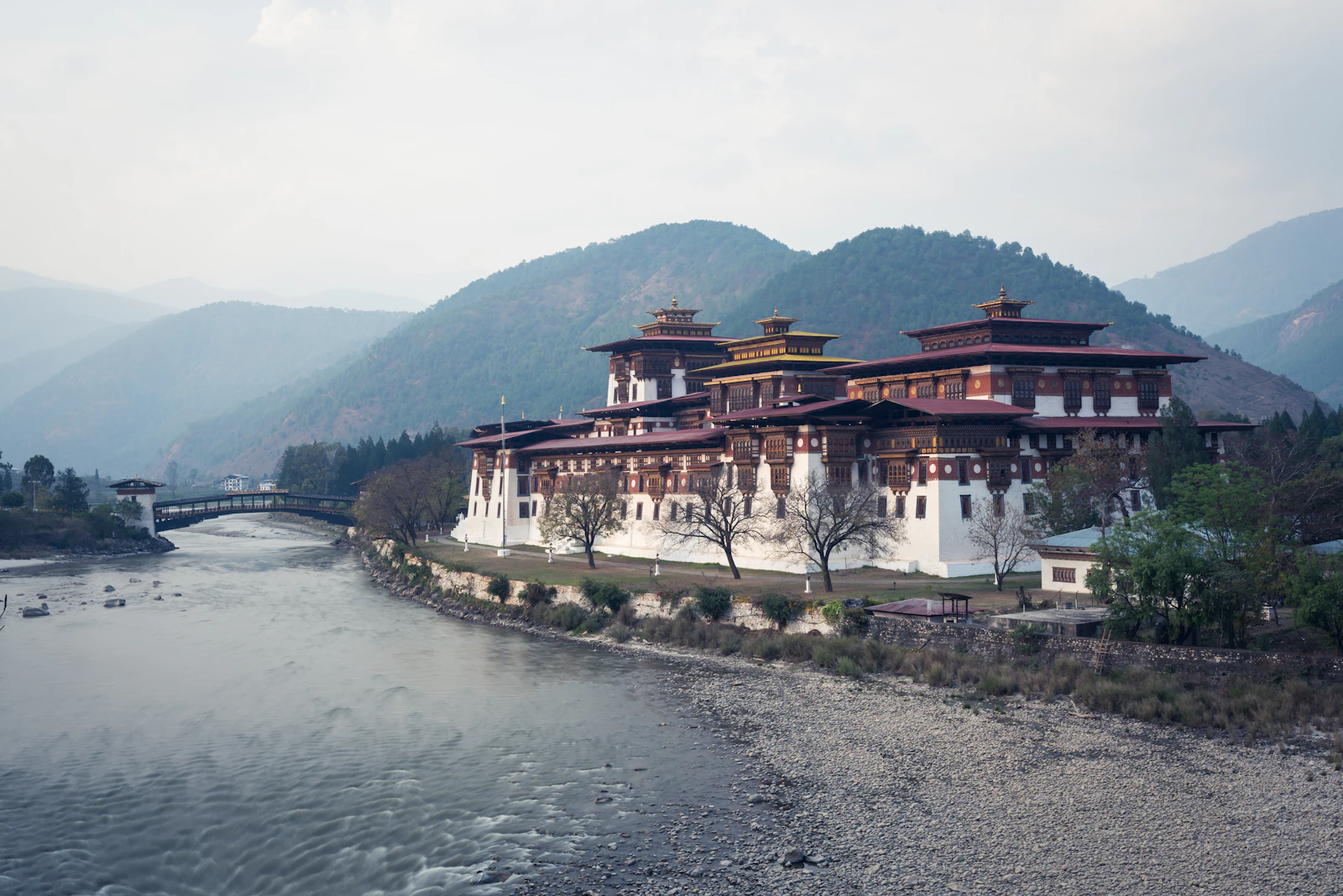 Punakha Dzong on the riverbank beside its covered bridge, forested hills rising behind under a soft sky