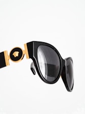 A close-up of stylish black sunglasses with gold detailing, featuring a decorative emblem. The design has a glossy finish and the lenses are tinted dark.