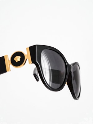 A close-up of stylish black sunglasses with gold detailing, featuring a decorative emblem. The design has a glossy finish and the lenses are tinted dark.
