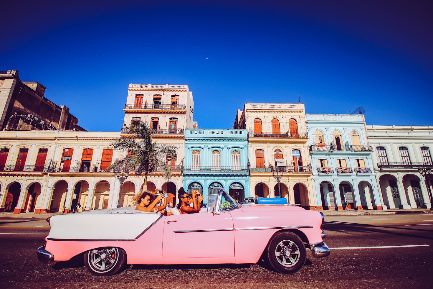 Delle persone in una auto storica rosa davanti agli edifici colorati di Cuba
