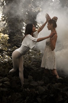Two women in a lush forest setting are engaged in an expressive, dramatic pose. One woman is wearing a white leotard, balancing on one foot, and reaching out towards the other woman, who is wearing a light dress and is standing amidst thick green foliage. A mist or smoke is visible in the background, adding an ethereal quality to the scene.