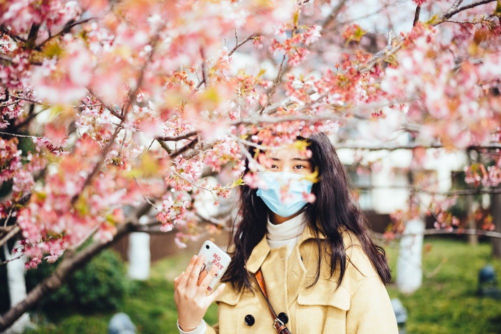 woman in brown coat with blue and white face mask holding white smartphone
