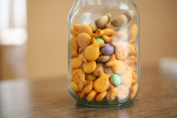A glass jar filled with goldfish-shaped crackers, round colorful candies, and striped wafer sticks. The jar is placed on a wooden table with a blurred, neutral background.