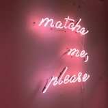 Neon pink cursive text on a wall reads 'matcha me, please' creating a warm and inviting glow. The text is affixed to a plain background, casting gentle shadows.