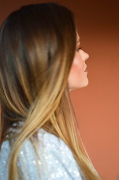 A person with long brown hair is shown in profile against a warm, muted orange background. The hair is smooth and falls over the shoulder, while the person is wearing a sparkly silver garment.