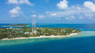 A small tropical island surrounded by clear blue waters and lush greenery. There is a tall telecommunications tower in the middle of the island, and several small boats are anchored near the shore. The sky is bright with a few scattered clouds, adding to the serene and picturesque setting.