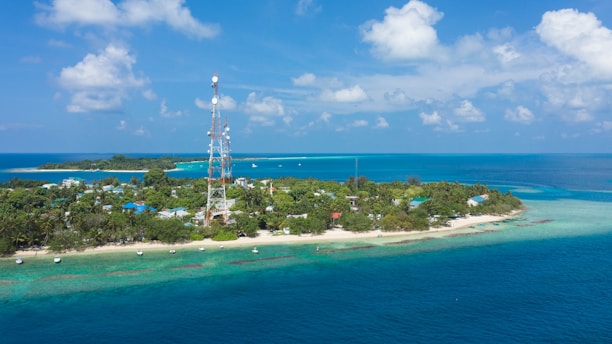 A small tropical island surrounded by clear blue waters and lush greenery. There is a tall telecommunications tower in the middle of the island, and several small boats are anchored near the shore. The sky is bright with a few scattered clouds, adding to the serene and picturesque setting.