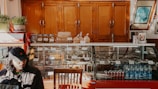 A cozy cafe interior features a wooden cabinet backdrop, a glass display case holding various pastries and snacks, and beverages including bottled water and Coca-Cola cans. To the left, a person in a black cap is seated by the counter. Plants and framed pictures add to the decor.