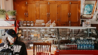 A cozy cafe interior features a wooden cabinet backdrop, a glass display case holding various pastries and snacks, and beverages including bottled water and Coca-Cola cans. To the left, a person in a black cap is seated by the counter. Plants and framed pictures add to the decor.