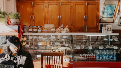 A cozy cafe interior features a wooden cabinet backdrop, a glass display case holding various pastries and snacks, and beverages including bottled water and Coca-Cola cans. To the left, a person in a black cap is seated by the counter. Plants and framed pictures add to the decor.