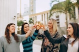 Four women are gathered outdoors, raising their glasses in a cheerful toast. They appear happy and engaged, smiling broadly, with a backdrop of palm trees and buildings.