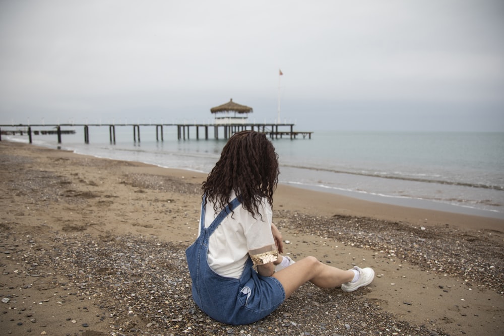 woman in white shirt and blue denim shorts sitting on beach shore during daytime