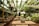 In an open-air setting with rustic wooden tables and benches in rows, small potted plants are placed on tables. The space is lush with greenery, featuring hanging and potted plants. Thatch-roofed structures and abundant natural light create a serene, garden-like ambiance.