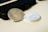 Two silver coins are placed on a light surface next to a black wallet or phone case. One coin displays an emblem of the United States with an eagle, while the other features a side profile of a bearded man with inscriptions.
