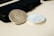 Two silver coins are placed on a light surface next to a black wallet or phone case. One coin displays an emblem of the United States with an eagle, while the other features a side profile of a bearded man with inscriptions.
