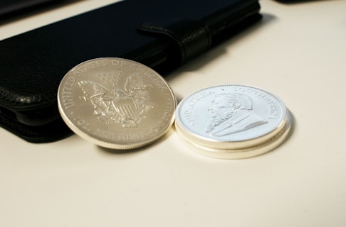 Two silver coins are placed on a light surface next to a black wallet or phone case. One coin displays an emblem of the United States with an eagle, while the other features a side profile of a bearded man with inscriptions.