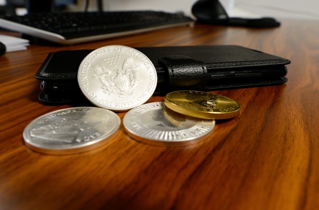 A black wallet is placed on a wooden desk with three silver coins and two gold coins laid out next to it. A keyboard is partially visible in the background, suggesting a workspace setting.
