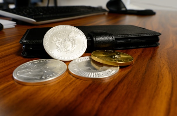 A black wallet is placed on a wooden desk with three silver coins and two gold coins laid out next to it. A keyboard is partially visible in the background, suggesting a workspace setting.