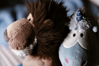 Two plush toys are positioned closely together. On the left is a brown lion with a fluffy mane and soft, round features. On the right is a blue unicorn with a fluffy mane, a small horn, and a friendly expression.