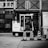 A black and white image depicting a construction or industrial area. A small utility vehicle is parked in front of a building with brick walls and metal structures. Several traffic cones with signs are aligned in front of the vehicle. The setting appears to be urban and utilitarian.