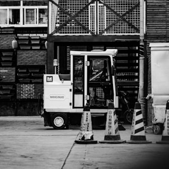 A black and white image depicting a construction or industrial area. A small utility vehicle is parked in front of a building with brick walls and metal structures. Several traffic cones with signs are aligned in front of the vehicle. The setting appears to be urban and utilitarian.