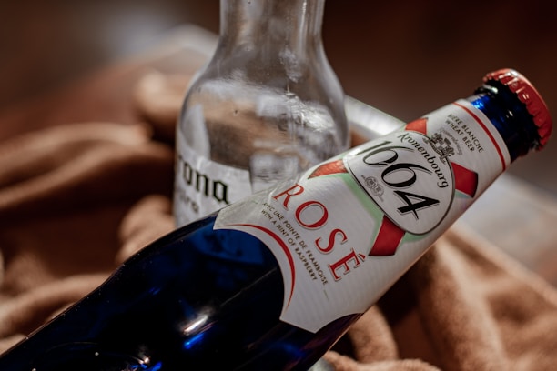 Two bottles are placed on a soft brown fabric. A clear glass bottle with 'Corona' branding is partially visible behind a blue bottle with a 'Kronenbourg 1664 Rosé' label, featuring red and white accents.