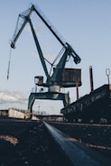 A large, industrial crane stands against the sky, with its metallic structure towering over the surroundings. The crane's arm extends upward, while a long rail or track runs along the ground towards the crane. The environment appears to be an industrial or shipping area with some buildings and clouds in the background.