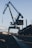 A large, industrial crane stands against the sky, with its metallic structure towering over the surroundings. The crane's arm extends upward, while a long rail or track runs along the ground towards the crane. The environment appears to be an industrial or shipping area with some buildings and clouds in the background.