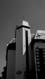 A tall, modern building with geometric architecture under a clear sky. The structure features a prominent vertical column and a triangular, sloped roof. Shadows create distinct lines on the facade, and there is a small sign reading 'TANGLIN PLACE' visible near the base.