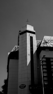 A tall, modern building with geometric architecture under a clear sky. The structure features a prominent vertical column and a triangular, sloped roof. Shadows create distinct lines on the facade, and there is a small sign reading 'TANGLIN PLACE' visible near the base.