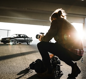 A person is kneeling on the ground with a camera, appearing to take a photograph of a car. The person is positioned in front of a bright light source, possibly the sun, which creates a dramatic backlighting effect. The car in the background is parked, and the setting appears to be a parking garage or an outdoor-covered area.